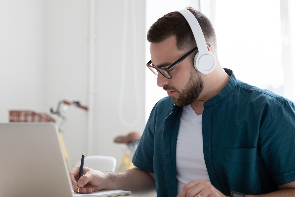 An adult learner in an online lesson, headphones on, taking notes beside a laptop